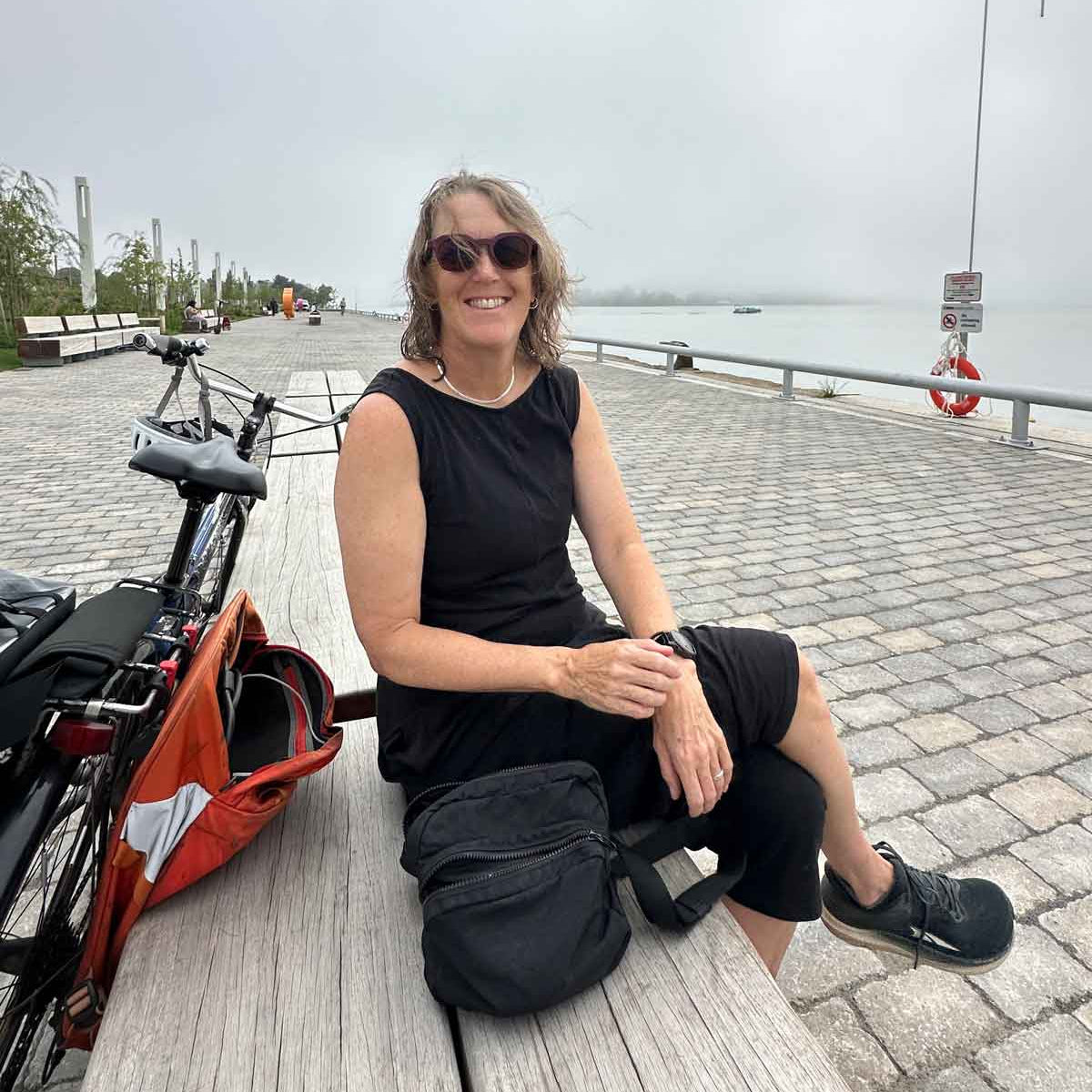 Allyson Woodrooffe wearing a black sleeveless top and black shorts sits on a wooden bench at a waterfront promenade, with a bicycle and an orange bag beside her.