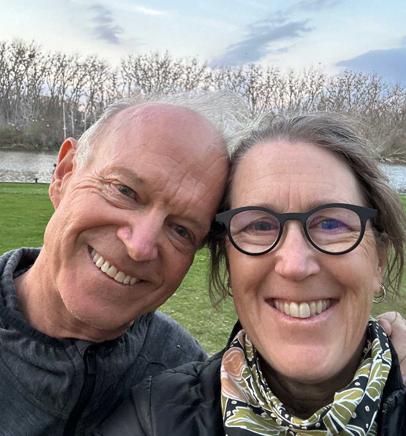 A close-up selfie of Philip Shepherd and Allyson Woodrooffe outdoors near a grassy field and trees. Allyson wears black-rimmed glasses and a patterned scarf, and Philip wears a dark zippered jacket.